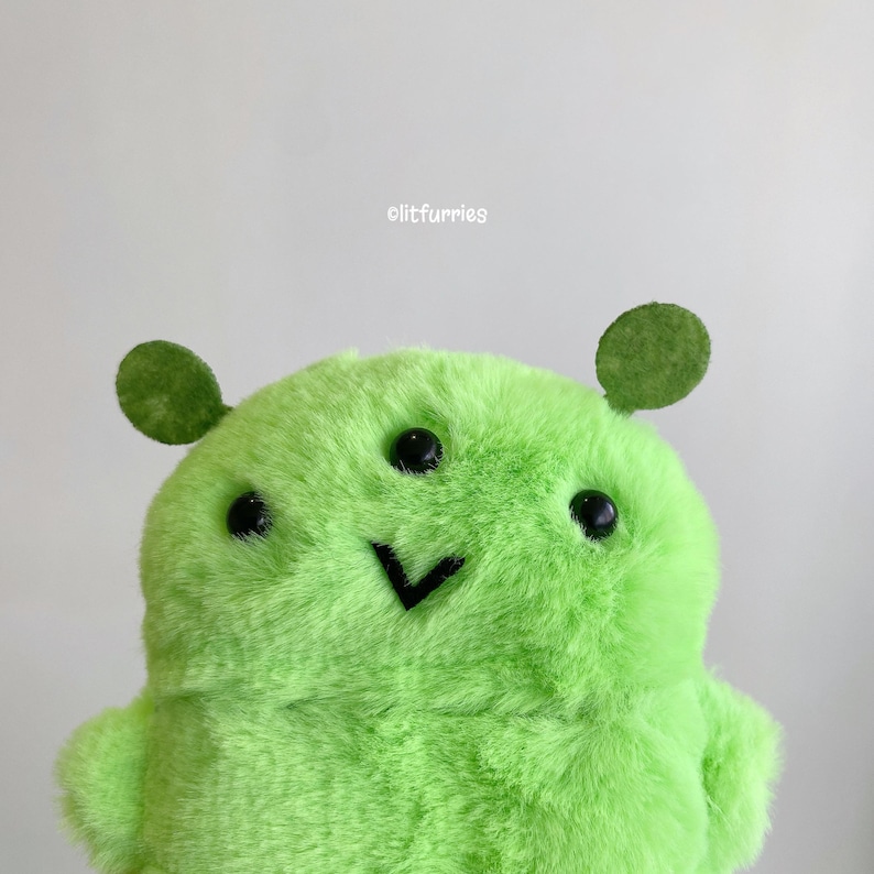 May include: A bright lime green plush toy with soft fur, featuring three black plastic eyes and a black V-shaped stitched mouth. The creature has two small, round, flat green ears on top and the text olitfurries printed above its head.