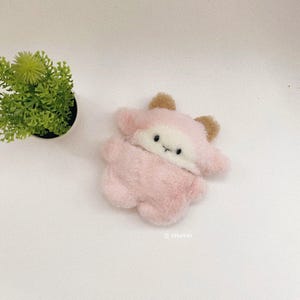 以下が含まれることがあります： 柔らかいピンクの毛並み、白い顔、小さな茶色の耳を持つ生き物の形をしたぬいぐるみ。このおもちゃには、2つの黒い目と小さな黒い刺繍の口があります。画像の右下隅にlitfurriesというテキストが表示されています。