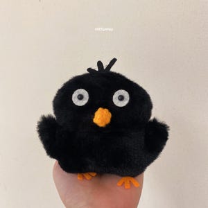 May include: A plush black bird with large white eyes, an orange beak, and orange feet. The bird has small wings and a tuft of feathers on its head. The bird is a stuffed animal.
