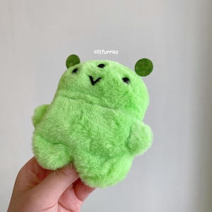 May include: A bright lime green plush toy with a soft, fuzzy texture, two small dark green circular ears, two black bead eyes, and a stitched black V-shaped mouth. The text litfurries is printed in white above the plush toy's head.