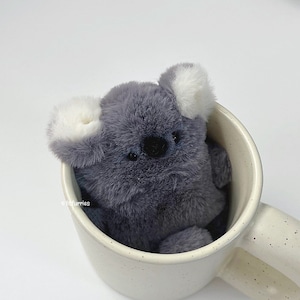 May include: A plush gray koala with white-tipped ears sits inside a speckled white mug. The koala has a black nose and small, dark eyes. The mug has a curved handle and is filled with the soft, furry toy.