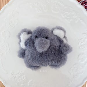 以下が含まれることがあります： 柔らかいグレーのフェイクファーで覆われた小さなゾウのぬいぐるみ。ゾウの耳の内側には白いアクセントがあり、2つの小さな黒いビーズの目と鼻が付いています。フィギュアの下には「littfurries」という文字が小さな白い文字で印刷されています。