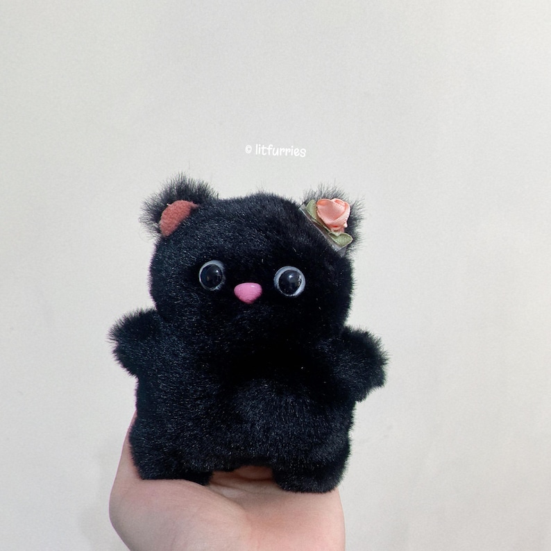 May include: A black plush toy bear with large eyes and a pink nose. The bear is wearing a small pink flower on its head.