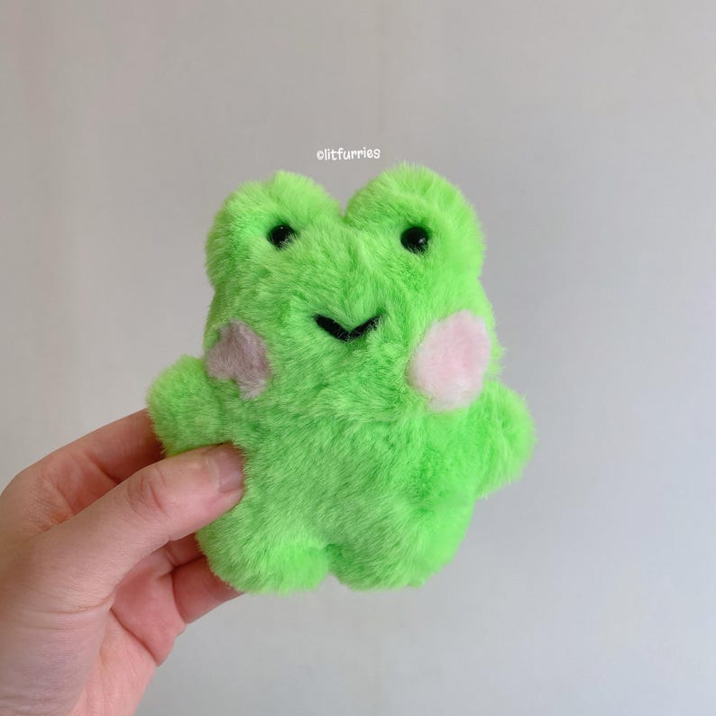 May include: A plush green frog toy with black eyes and a stitched black mouth. The frog has pink blush spots on its cheeks and is made of soft, fuzzy material. The text "clitfurries" is visible in the upper right corner.