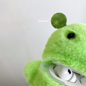 May include: A bright lime green fuzzy plush case for wireless earbuds. The case features a round ear-like detail on top, a single black eye, and the text "litfurries" printed in white. The white charging case is visible inside the plush cover.