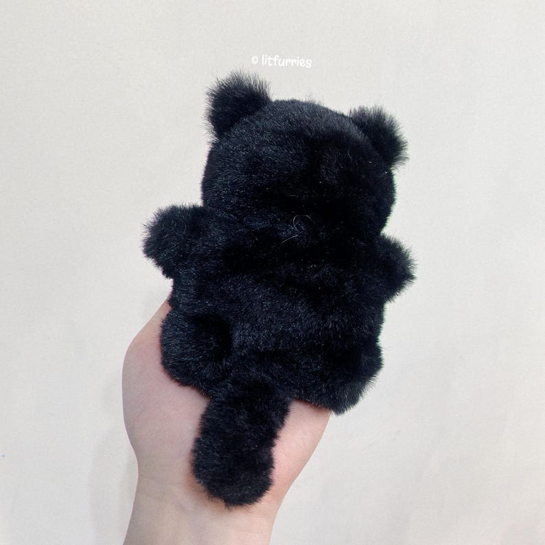 May include: Black plush cat toy with a long tail. The cat is sitting up with its back to the camera.