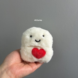 May include: A white plush toy with a soft, fuzzy texture, featuring two small black eyes, a stitched black smile, and a bright red heart patch on the front. The text litfurries is printed in small white letters above the plush object.
