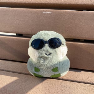 May include: A light green plush toy shaped like a blob, with white patches and green details. It wears black sunglasses and has a stitched black mouth. The background features the text "olitfurries".
