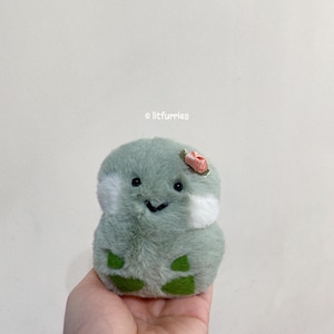 May include: A plush green frog toy with a small pink flower on its head. The frog has a white belly and a friendly face.