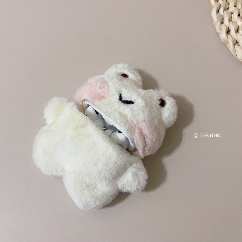 Op de afbeelding: Wit pluche etui in de vorm van een kikker voor draadloze oordopjes. Het etui heeft roze accenten en een glimlachend gezicht.