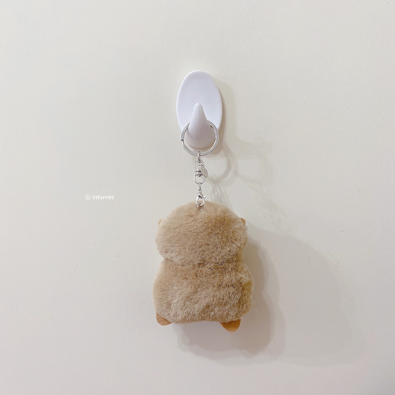 K&ouml;nnte beinhalten: Ein brauner, flauschiger Schl&uuml;sselanh&auml;nger mit einer silbernen Kette, die an einem wei&szlig;en Haken an einer wei&szlig;en Wand h&auml;ngt.