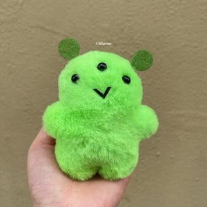 Può includere: Un giocattolo di peluche verde con un viso sorridente e tre occhi. Il giocattolo ha una consistenza pelosa e ha la forma di una creatura con grandi orecchie.