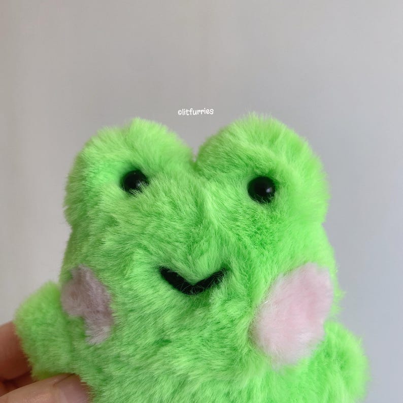 May include: A plush green frog with black eyes and a smiling mouth. The frog has light pink blush marks on its cheeks. The text "olitfurries" is visible on the top right of the image.