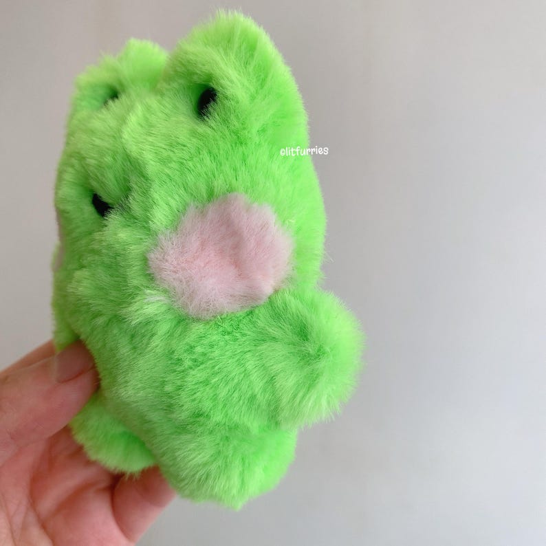 May include: A plush green frog with a pink heart-shaped patch on its chest. The frog has black eyes and is made of soft, fuzzy material. The text "olitfurries" is visible on the side of the frog.