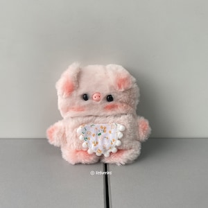 May include: A plush pink pig-shaped item with black eyes, a pink snout, and blush-colored cheeks. It has a small, white bib with a yellow floral pattern. The item is fluffy and has small, rounded ears.