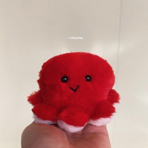 May include: A red plush octopus with a white smile and black eyes. The octopus is sitting on a hand.