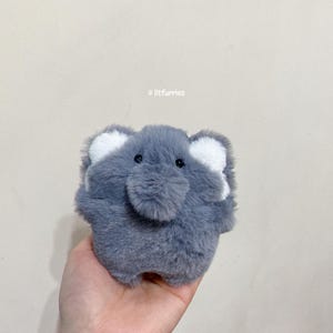 May include: A small gray plush toy shaped like an elephant with white ears. The toy has black eyes and a fluffy texture.