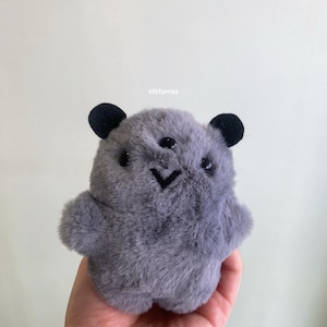 Può includere: Un peluche grigio con occhi neri a bottone, orecchie nere e bocca nera cucita. Il giocattolo ha una consistenza soffice e un corpo arrotondato. Il giocattolo è tenuto in una mano.