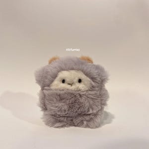 以下が含まれることがあります： ふわふわとした外観とクリーム色の顔を持つ、グレーのぬいぐるみ。小さな黒い目と縫い付けられた笑顔があります。頭の上部には、2つの小さなタン色の耳があります。「clitfurries」というテキストが上部に表示されています。
