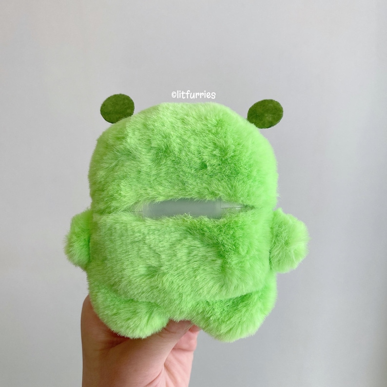 May include: A bright lime green plush case for wireless earbuds with a soft, fuzzy texture. The case features two small, rounded green ears on top and a horizontal cutout in the center. The text olitfurries is printed in white above the cutout.