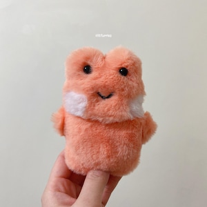 May include: A plush orange frog with white accents and black eyes. The frog is smiling and has a fluffy texture.