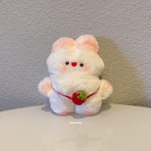 以下が含まれることがあります： ピンクの耳と赤い緑のイチゴがついた白いぬいぐるみウサギ。ウサギは腰に赤いリボンをしています。