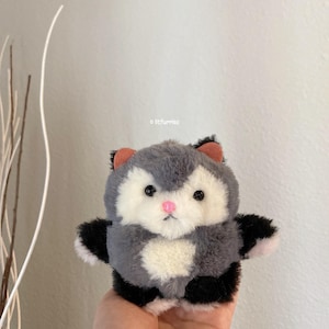 May include: A round plush toy featuring grey and white faux fur, black paws, and small peach-coloured ears. The creature has black plastic eyes, a small pink nose, and the text litfurries printed in small black letters above its head.
