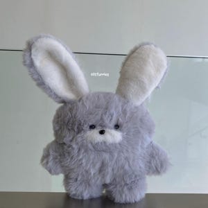 Peut inclure: Une peluche lapin grise avec de longues oreilles doublées de blanc et un corps duveteux. Le jouet en peluche a un petit nez noir et des yeux foncés. Le texte "clitfurries" est visible sur l'image.