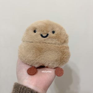 May include: A plush, tan-colored creature with a smiling face and small, round, brown feet. The creature has black embroidered eyes and a curved black embroidered mouth. The plush toy is held in a hand.