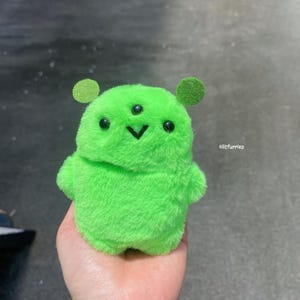 May include: A plush green alien toy with three eyes and two small ear-like circles. The alien has a small black v-shaped mouth and small arms. The toy is held in a hand. The text "olitfurries" is in the image.