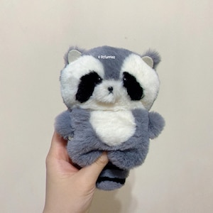 May include: A plush raccoon toy with soft grey and white fur, featuring black eye patches, a small black nose, and a striped tail. The text litfurries is printed in small white letters near the top of the raccoon's head.