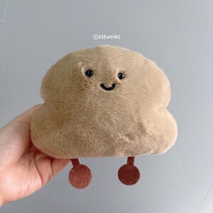 May include: A light brown, cloud-shaped plush toy with a smiling face, black eyes, and a curved mouth. It has two small, brown, rounded feet. The text "@litfurries" is visible in the upper right corner of the image.