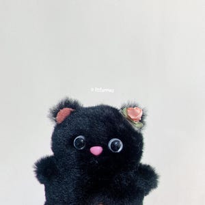 May include: A black plush toy with large eyes and a pink nose. The toy is wearing a small pink flower on its head.