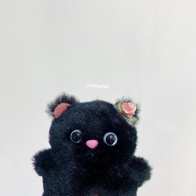May include: A black plush toy with large eyes and a pink nose. The toy is wearing a small pink flower on its head.