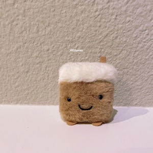 May include: A brown and white plush toy shaped like a cup of coffee with a smiling face. The toy has a white fluffy top and brown fur on the bottom. The toy is sitting on a white surface.