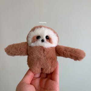 Puede incluir: Un juguete de peluche de perezoso hecho a mano con los brazos extendidos. El perezoso tiene pelaje marrón con una cara blanca y marrón y ojos negros grandes. El texto "clitfurries" es visible en la parte superior de la imagen.