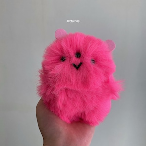 May include: A bright pink, fluffy plush toy with a cartoonish face and small ears. The toy has a round body and is made of soft, furry material. The face features three black dots for eyes and a black chevron for a mouth. The text "elitfurries" is in the top left corner.