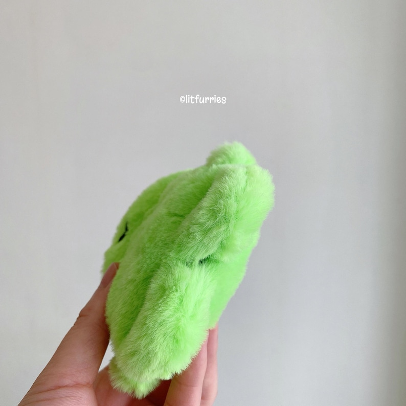 May include: A bright lime green plush accessory with a soft, fuzzy texture. The item features a rounded, irregular shape and includes the text "litfurries" printed in small white letters near the top edge of the object against a plain background.