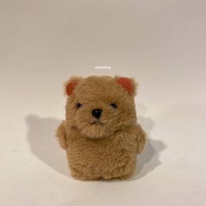Può includere: Un orsacchiotto marrone con orecchie arancioni e un naso nero. L'orso ha una consistenza morbida e pelosa e un'espressione leggermente triste. L'orso è un oggetto fatto a mano.