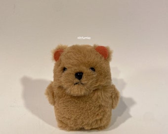 May include: A small, plush, light brown bear toy with soft, fuzzy fur. It features a black nose, dark eyes, and small, rounded ears with orange-colored inner accents. The text 'olitfurries' is printed in small white letters above the bear's head.