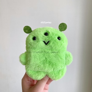 May include: A plush, lime green alien-like creature with three black eyes, two green circular antennae, and small arms and legs. The creature has a black stitched smile. The text "elitfurries" is in the upper right corner.