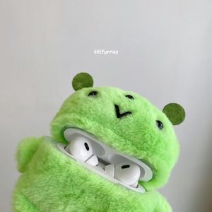 May include: A bright lime green plush fuzzy case for wireless earbuds, featuring two small round ears, a stitched black V-shaped mouth, and two black eyes. The case is open, revealing white earbuds inside. The text "olitfurries" is printed above.