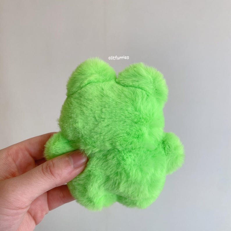 May include: A bright green plush frog toy with a soft, furry texture. The frog has a rounded body, four small limbs, and two large, rounded eyes. The toy is held in a hand against a plain white background.