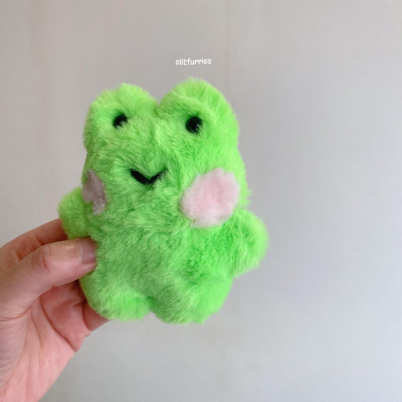May include: A plush green frog toy with black eyes and a stitched black mouth. The frog has pink blush marks on its cheeks and a soft, furry texture. The text "clitfurries" is visible in the upper right corner.