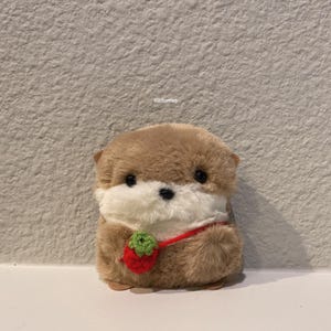 May include: A small, plush, tan and white otter toy with black eyes and a black nose. The otter wears a red crocheted strawberry-shaped bag with a green stem on a thin red strap. The text clitfurries is printed in small white letters above the toy.