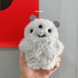 May include: A small, round, light gray plush creature with long, soft fur. It features three black bead eyes, two small dark gray rounded ears, and a stitched mouth. The text litfurries is visible above the creature's head in white font.