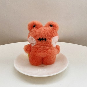 Puede incluir: Un juguete de peluche con forma de rana naranja con detalles cosidos en negro para los ojos y la boca, y acentos blancos en las mejillas. La rana está sentada en un plato blanco. El juguete es un artículo lindo y hecho a mano.