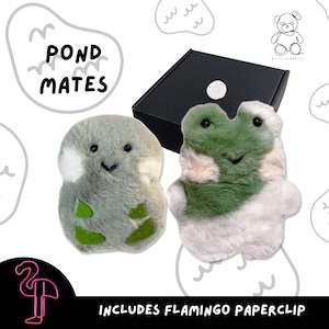 May include: Two plush toys, one grey and one green, with a black box. The grey toy has white and green accents, while the green toy has pink and white accents. The text "POND MATES" is above the toys, and the text "INCLUDES FLAMINGO PAPERCLIP" is below.