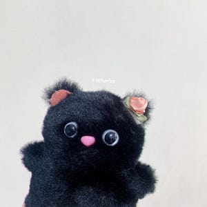 May include: Black plush toy with a pink nose and large black eyes. The toy has a pink flower on its head and is made to look like a cat.