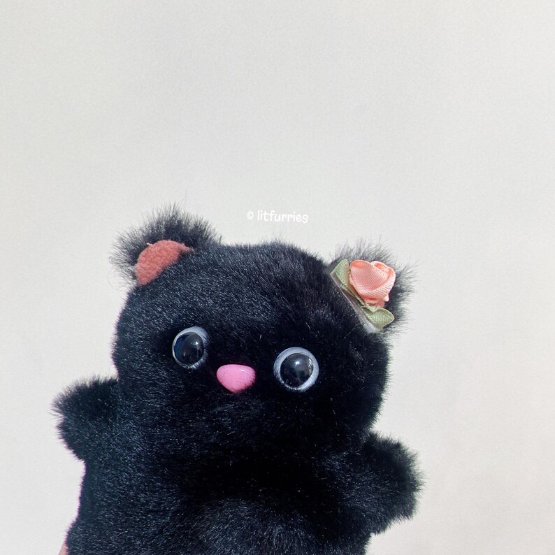 May include: Black plush toy with a pink nose and large black eyes. The toy has a pink flower on its head and is made to look like a cat.